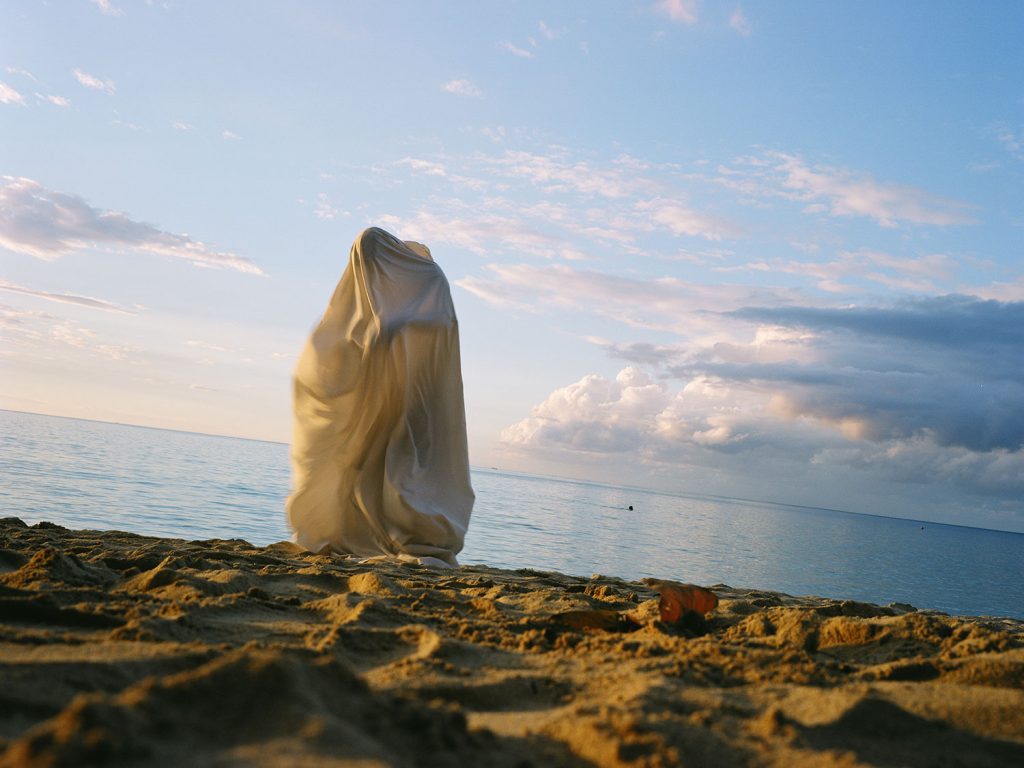 personin white dress standing on beach, looking out at ocean under clear blue sky.