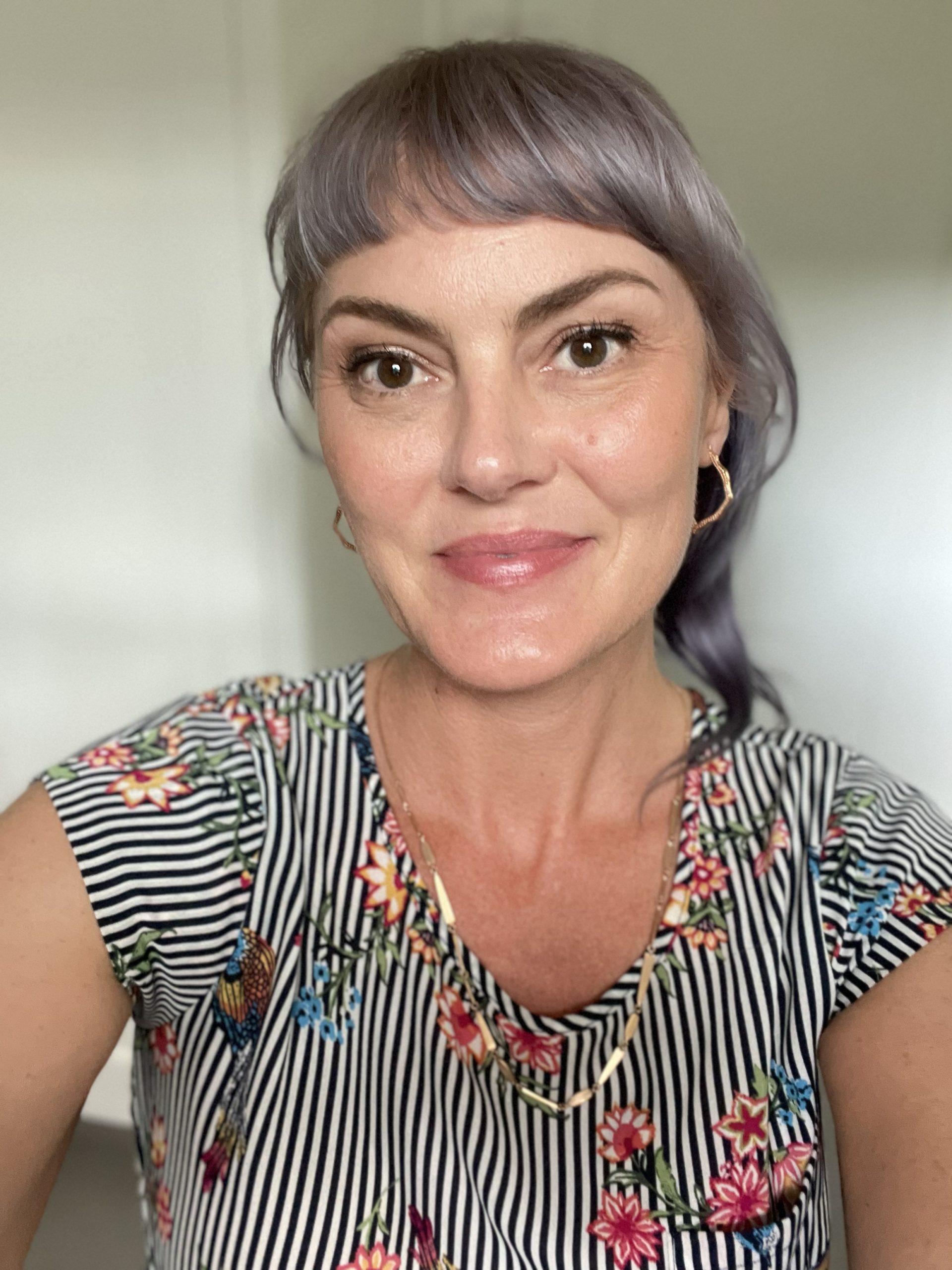 Ami Mariscal smiles at the camera, wearing a striped floral shirt and gold hoop earrings, with light purple hair.