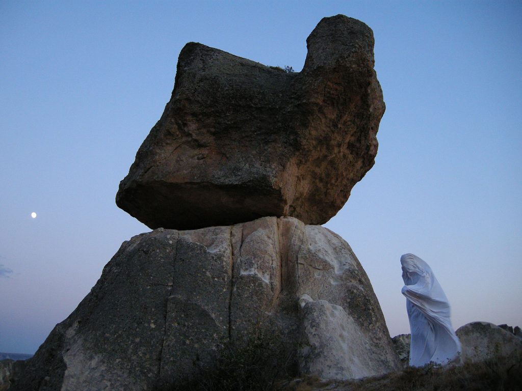 A person in a white robe standing next to a large rock in a serene landscape.