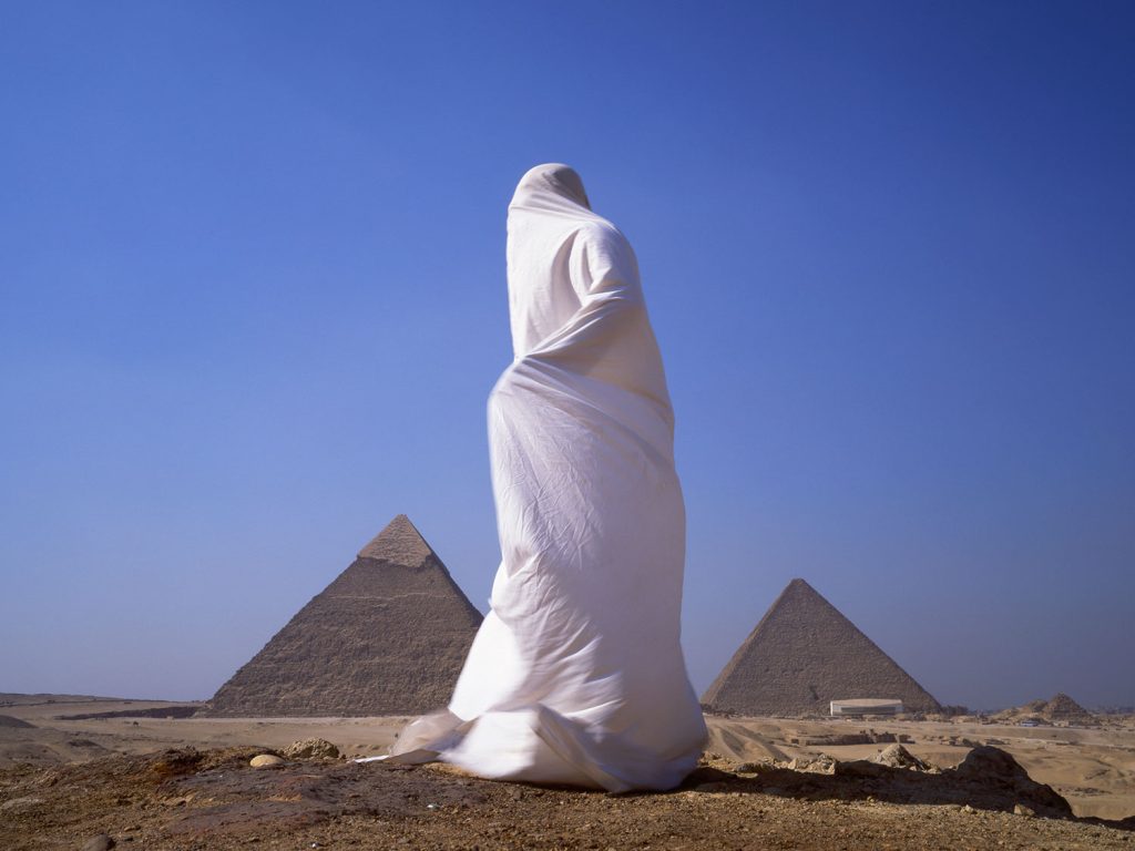 A person wearing white robe in a shroud stands in front of the pyramids.