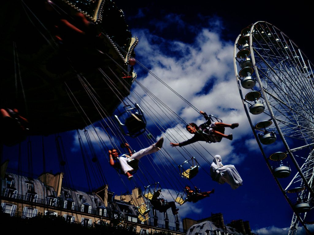 A ferris wheel with colorful gondolas and people enjoying swings at an amusement park on a sunny day.