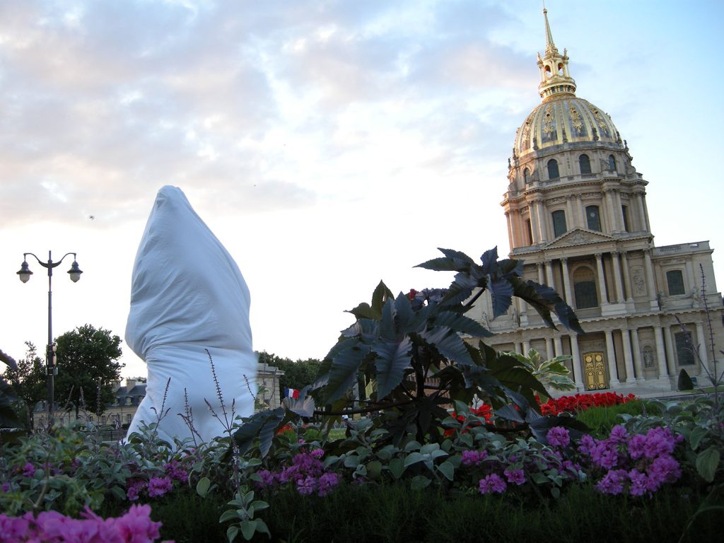 A white umbrella rests in a flower ground, complementing the personwearing white cloth standing outside near the white minar.