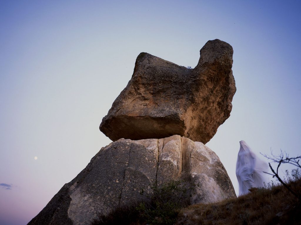 A person standing white robe draped on top of a large rock formation, overlooking a vast landscape.