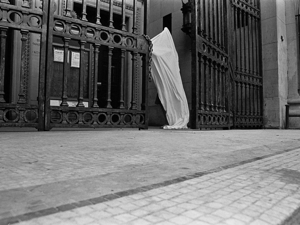 A person standing draped white robe  in front of a gate, captured in black and white.