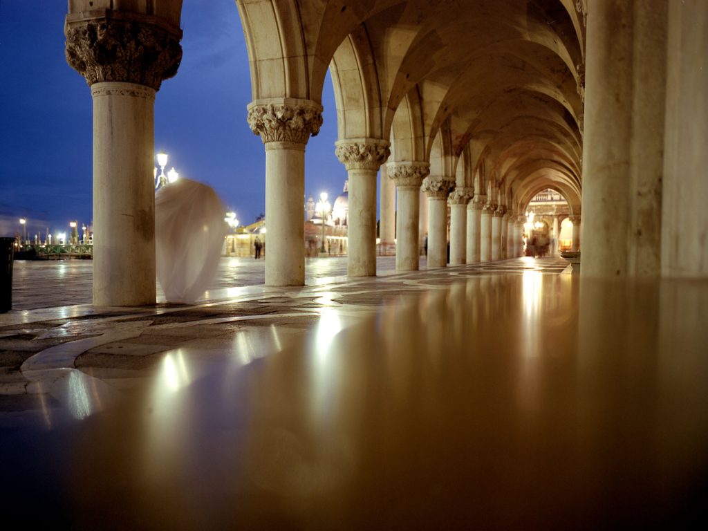 A person wearing white robe standing a long walkway with illuminated pillars and arched doorway at night.