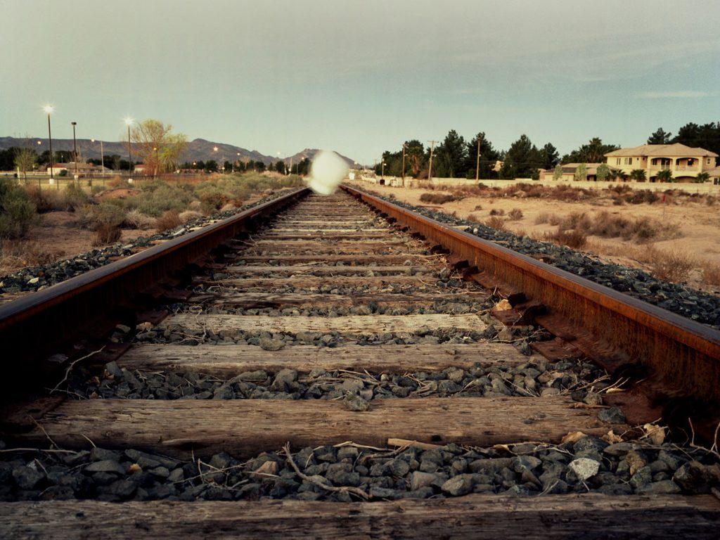 A person draped with white cloth resting on a train track, creating a sense of anticipation and potential danger.