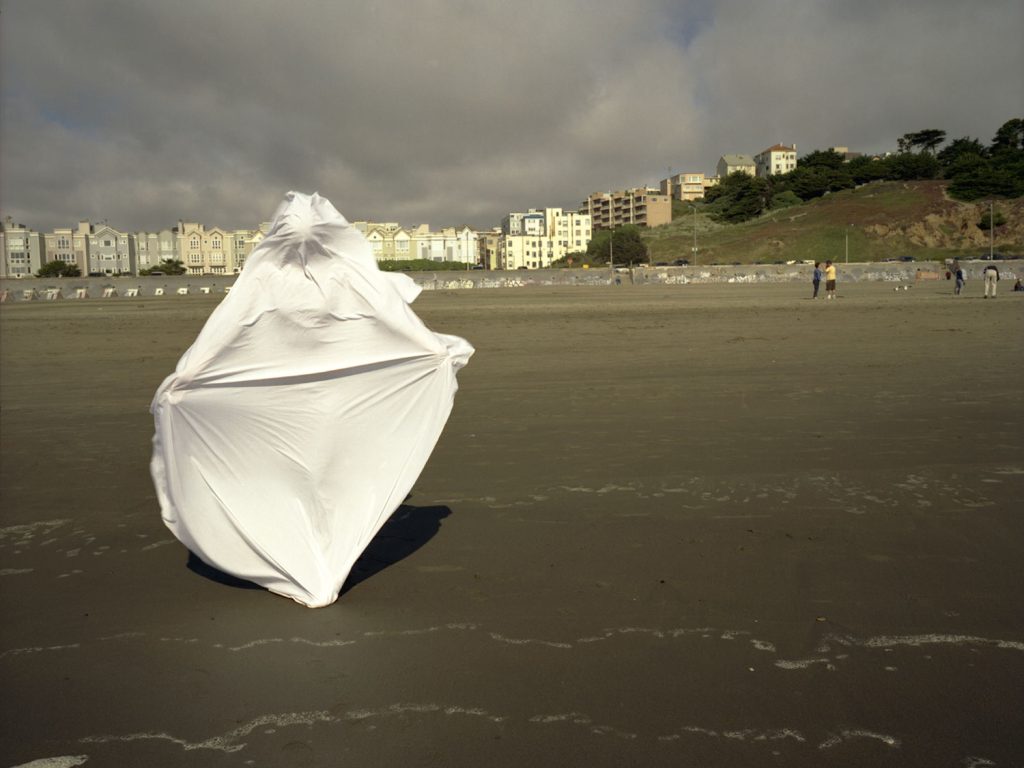 A person in a white robe standing on a beach, looking out at the ocean waves under a clear cloudy sky.