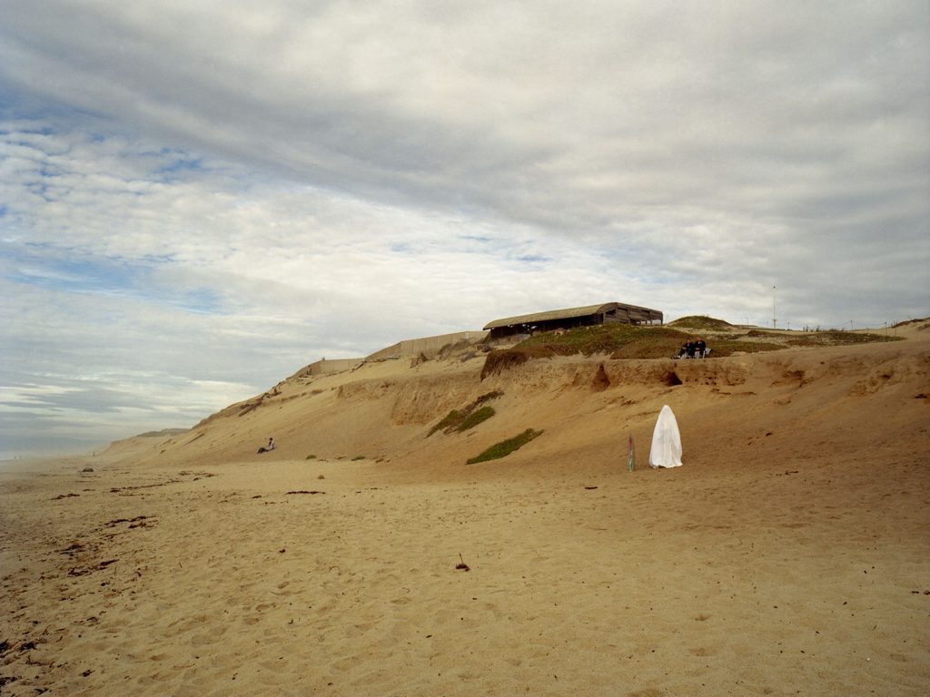 A person wearing white cloth standing on sandy beach, providing shade on a sunny day.